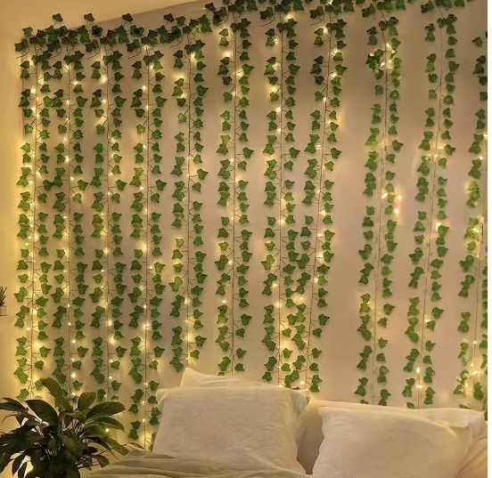Warm white fairy string lights woven through artificial ivy vine garlands draped across a bedroom wall above a white bed