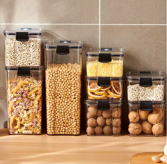 Set of clear airtight food storage containers with black locking lids stacked on a kitchen counter filled with pasta, grains, nuts and dried fruits